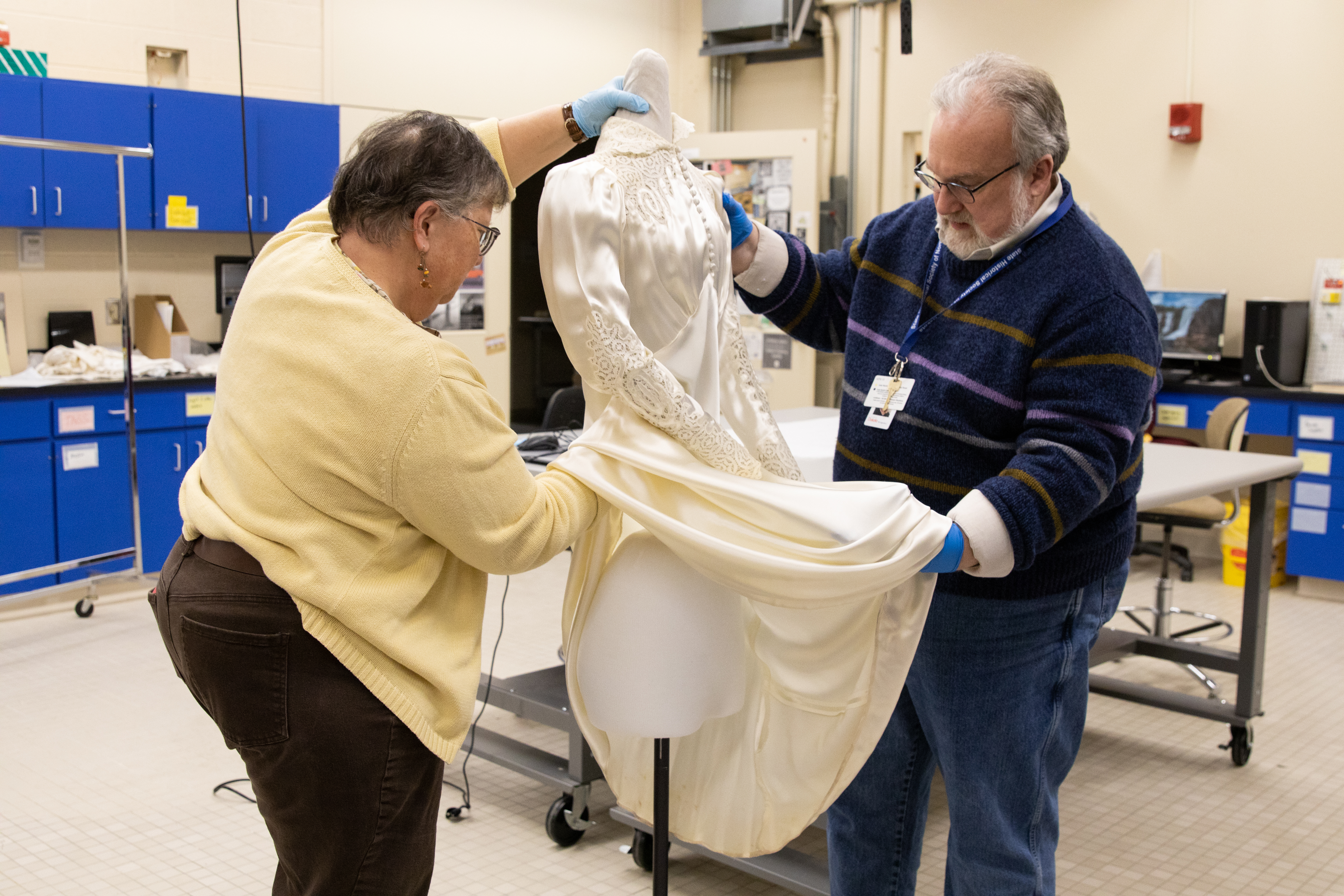 A man and woman dress a mannequin in an old wedding dress