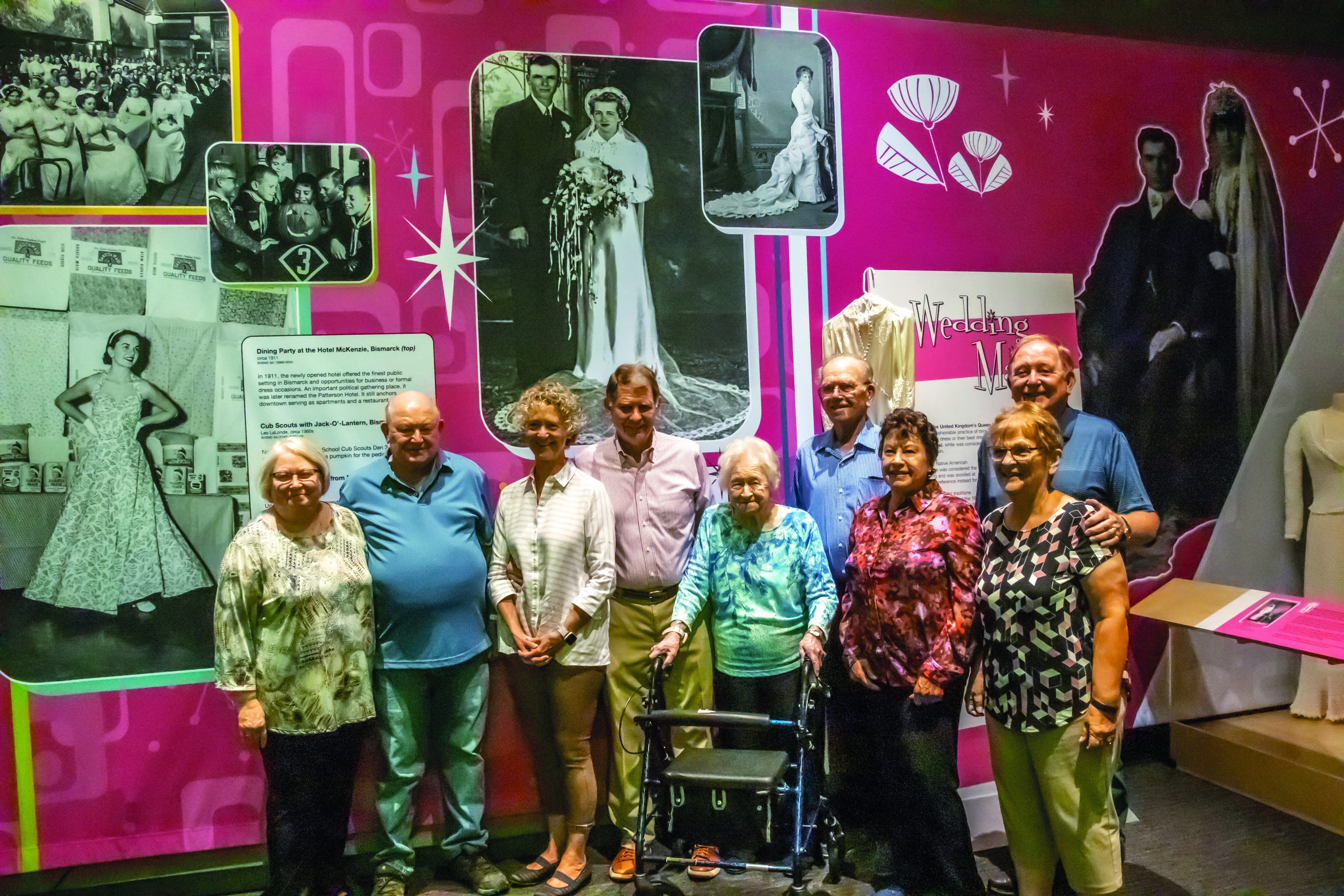 A old woman with a walker stands with four of her children and their spouses in front of her wedding gown and a photo of her and her late husband on their wedding day that are on display for a museum exhibit.