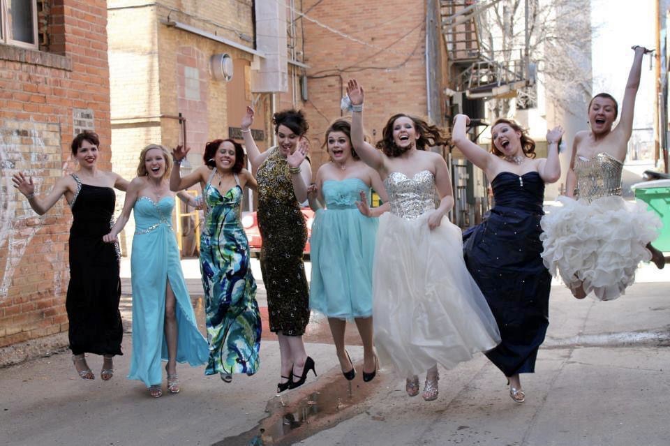 8 girls wearing prom dresses jump in the air to pose for a photo between brick buildings