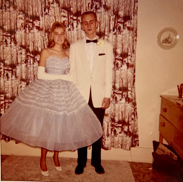 A young woman wearing a poofy blue dress with white shoes and above the elbow gloves stands with her arm linked with a young man in a white suite coat with a black bowtie, pants, shoes, and handkerchief in the breast pocket of the coat